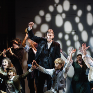 An image of our first production of Charles Dickens' 'A Christmas Carol'. The stage is full of musicians and people singing with the central character of the play holding one arm to the air in jubilation.