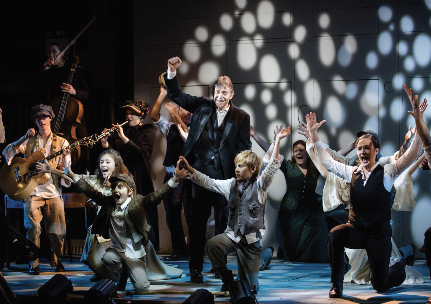 An image of our first production of Charles Dickens' 'A Christmas Carol'. The stage is full of musicians and people singing with the central character of the play holding one arm to the air in jubilation.